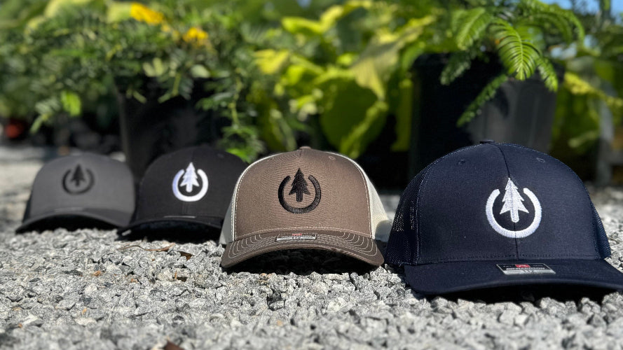 Four baseball caps with different designs on a gravel surface with greenery in the background